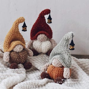 May include: Three plush gnomes with long, pointed hats and round bodies. Each gnome has a different colored hat: mustard yellow, burgundy red, and light gray. They each hold a miniature black lantern with a yellow glow. The gnomes are sitting on a soft, white blanket.
