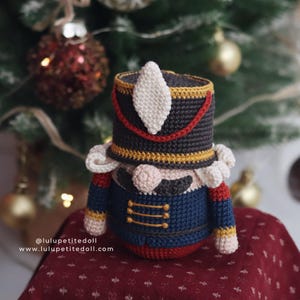 May include: A handmade crochet Nutcracker doll in a festive Christmas setting. The doll features a black hat with red and gold trim, a blue jacket, and a rosy-cheeked face. The background includes a Christmas tree and ornaments.