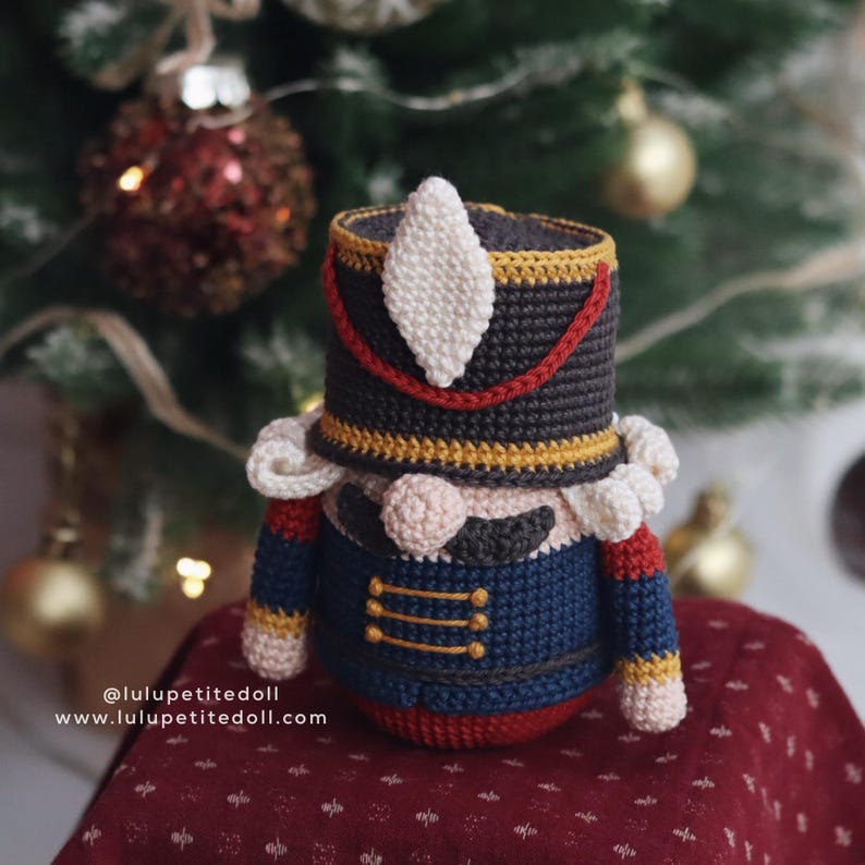 May include: A handmade crochet Nutcracker doll in a festive Christmas setting. The doll features a black hat with red and gold trim, a blue jacket, and a rosy-cheeked face. The background includes a Christmas tree and ornaments.