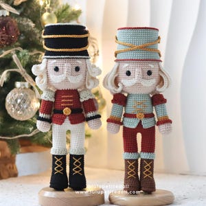 Puede incluir: Dos muñecas cascanueces de crochet, una en rojo y blanco y la otra en azul y blanco, de pie sobre bases de madera. Ambas muñecas tienen un ribete dorado en sus sombreros y un botón dorado en sus chaquetas.