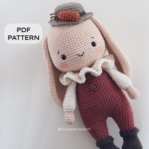 May include: A crocheted bunny doll wearing a brown hat with a carrot on top, a white shirt with a ruffled collar, brown pants, and brown boots. The doll has a smiling face with black eyes and a pink blush.