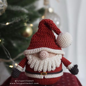 May include: A handmade crochet Santa Claus doll. The doll features a red hat with a white pom-pom, a white beard, and a red suit with a black belt. The doll's arms and gloves are black. The doll is sitting on a red surface. The text on the image reads: @lulupetitedoll www.lulupetitedoll.com