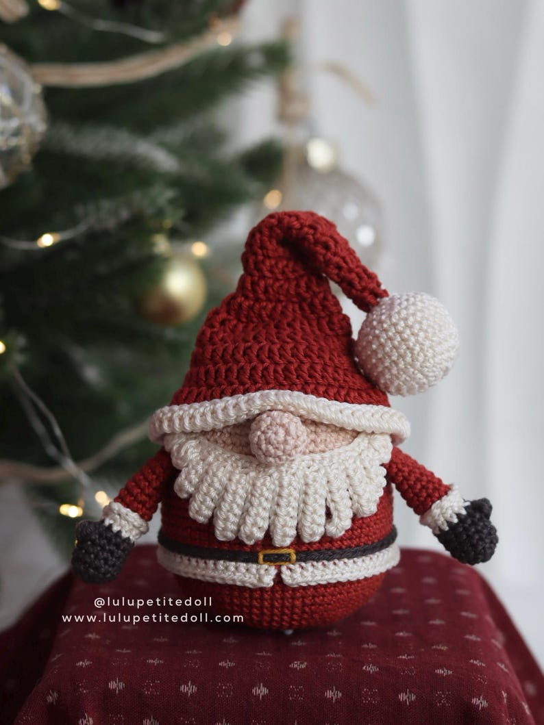 May include: A handmade crochet Santa Claus doll. The doll features a red hat with a white pom-pom, a white beard, and a red suit with a black belt. The doll's arms and gloves are black. The doll is sitting on a red surface. The text on the image reads: @lulupetitedoll www.lulupetitedoll.com