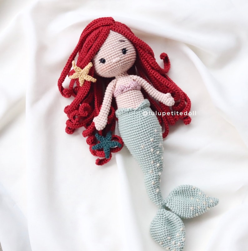 May include: A crocheted mermaid doll with red hair, a pink top, and a teal tail. The doll is holding a small teal starfish in its hand. The doll is sitting on a white surface.