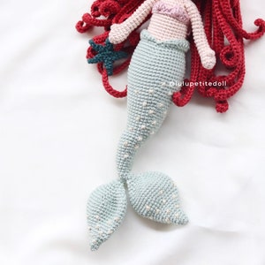 May include: A crocheted mermaid doll with a light blue tail, red hair, and a pink top. The doll is holding a dark blue starfish.