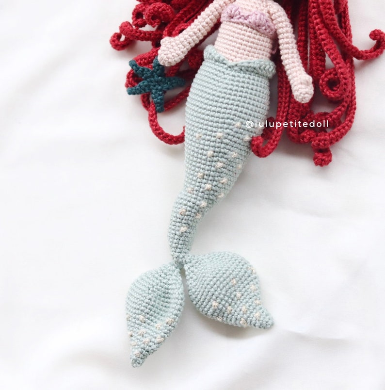 May include: A crocheted mermaid doll with a light blue tail, red hair, and a pink top. The doll is holding a dark blue starfish.