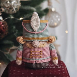 May include: A handmade crochet nutcracker doll in shades of pink, green, and brown. The doll has a detailed design with a hat, jacket, and decorative accents. The doll is approximately 6 inches tall and is a festive holiday decoration.