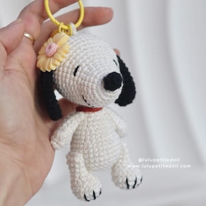 May include: A handmade white and black Snoopy keychain with a yellow flower and red collar. The crochet Snoopy has black ears and a black nose. The keychain is attached to a yellow ring.