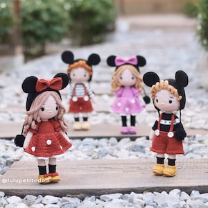 May include: Four crocheted dolls dressed in red, pink, and black outfits. Each doll has a black Mickey Mouse-style hat with large ears. The dolls are standing on a wooden surface with small rocks.