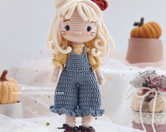 May include: A crocheted doll with blonde hair, wearing a blue overall dress and red shoes. The doll has cat ears and a red bow on its head. The doll is standing on a white surface with gold stars.