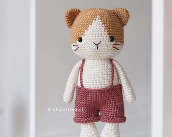 May include: A crocheted cat doll wearing a pair of red overalls. The cat has a white face with brown ears and a brown patch on its head. The doll is standing on a white shelf. @luluptitedoll