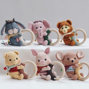 Puede incluir: Seis juguetes de peluche de crochet con anillos de madera. Los juguetes incluyen un burro, un elefante, un tigre, un oso, un cerdo y un animal marrón con orejas grandes. Todos los juguetes llevan bufandas y sostienen los anillos de madera.