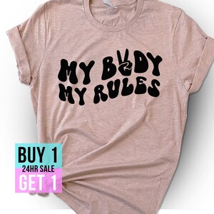 May include: A light brown t-shirt with the text "MY BODY MY RULES" in black, stylized lettering. The text includes a peace sign symbol. The shirt is on a white background. The text "BUY 1 GET 1 24HR SALE" is in a pink and blue box in the lower left corner of the image.