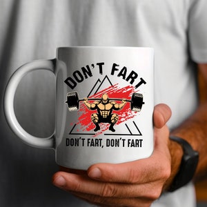 May include: White ceramic mug with a black handle featuring the text "DON'T FART" above a weightlifter graphic. The design includes a barbell and a red splatter effect, with the phrase "DON'T FART, DON'T FART" below.