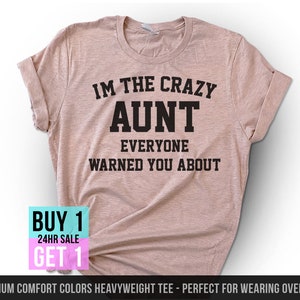 May include: A light pink t-shirt with the text "IM THE CRAZY AUNT EVERYONE WARNED YOU ABOUT" in black. The shirt has rolled-up sleeves and a crew neck. A "BUY 1 GET 1" sale graphic is in the lower left corner.