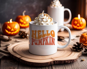 May include: White ceramic mug with the text "HELLO PUMPKIN" in orange and yellow letters. The mug is filled with a creamy beverage topped with whipped cream. The mug is sitting on a wooden slice with small pumpkins and pine cones around it.