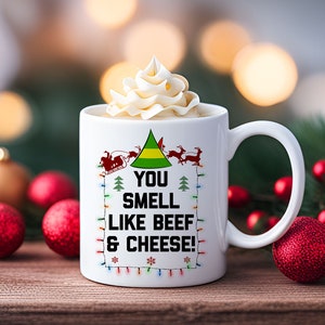 May include: White ceramic mug with a green Christmas tree and the text "You smell like beef & cheese!" in a red frame. The mug is filled with a white creamy beverage and is surrounded by red and gold Christmas ornaments.