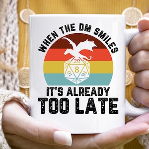 May include: White ceramic mug with a retro-style graphic of a dragon and a 20-sided die. The text reads "When the DM smiles it's already too late".