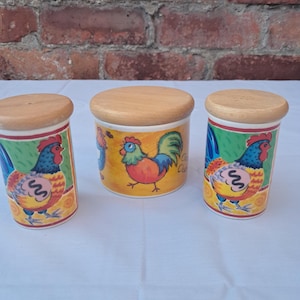 May include: Three ceramic canisters with light wood lids. Two tall, narrow canisters and one shorter, wider canister feature colourful illustrations of roosters with blue, red, and yellow feathers against green and yellow backgrounds.