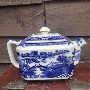 May include: A rectangular ceramic teapot featuring a blue and white willow pattern. The design depicts a landscape with a bridge, trees, and pagodas. It includes a matching lid with a small knob, a side handle, and a spout, all finished with gold trim.