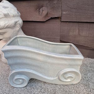 May include: A light blue ceramic planter with a smooth, glossy finish. The vessel features a rectangular opening with curved, wave-like sides and prominent scroll-shaped details at the base on both the left and right ends of the structure.