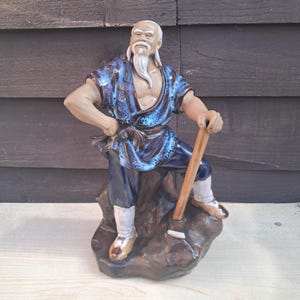 May include: A ceramic figurine of an elderly man in a blue and brown robe, holding a wooden staff. The figure has a long white beard and is standing on a brown rock base. The figurine is approximately 25 cm tall.
