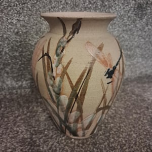 May include: A beige ceramic vase with a wide body and a narrow neck. The vase is decorated with painted dragonflies, reeds, and other plant life in shades of brown, green, and pink. The vase has a smooth, matte finish.