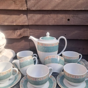 May include: A white porcelain tea set featuring a teal band with a geometric orange and brown pattern. The set includes a teapot with a lid, a creamer, a sugar bowl, and four teacups with matching saucers, all decorated with the same teal motif.