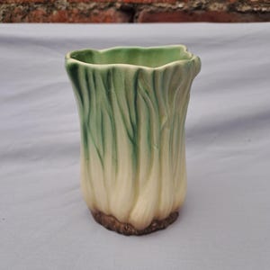 May include: A ceramic vase shaped like a head of bok choy or napa cabbage. The vessel features a textured, ribbed exterior with a gradient of creamy white at the base, transitioning into vibrant green hues towards the scalloped, open top rim.