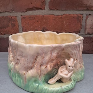 May include: A ceramic planter shaped like a tree stump, with a light pink and brown exterior. The planter has a light green base and a small, seated figurine. The interior is a light cream colour.