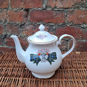 May include: A white ceramic teapot with a lid, gold trim, and handle. The teapot is decorated with a floral design on the lid and a tartan ribbon with a crest on the body. The teapot is placed on a woven wicker surface.