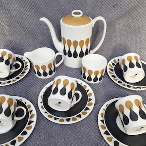 May include: A vintage tea set with a white teapot with a gold lid, a creamer, cups, and saucers. The set has a black and gold teardrop pattern. The saucers and cups are black with a gold and white trim.