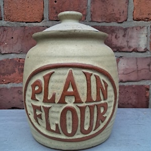 May include: A beige ceramic flour jar with a lid, displaying the words "PLAIN FLOUR" in a brown oval. The jar is set against a red brick wall, giving a vintage feel. The jar is designed for storing flour.
