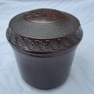 May include: A dark brown cylindrical container with a lid featuring a mottled finish, a textured band, and a decorative border of embossed leaves. The word Tobacco is embossed on the centre of the lid in a script font. The base is smooth and solid.