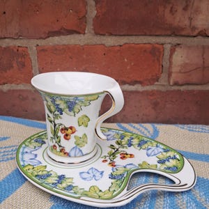 May include: A white teacup and saucer set with a botanical design of green leaves, blue berries, and orange fruit. The cup has a curved handle with gold trim, and the saucer has a unique handle shape. The set is placed on a blue and white patterned surface.