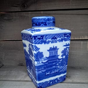 May include: A blue and white ceramic tea caddy with a lid. The caddy features a traditional Willow pattern design with a pagoda, trees, and a bridge. The square-shaped container is decorated with detailed landscape scenes and floral patterns.