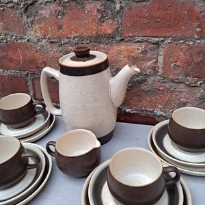 May include: A vintage ceramic coffee set. The set includes a cream-coloured coffee pot with a brown rim and handle, cups, saucers, and a creamer. The set has a speckled finish and a brown and cream colour scheme.