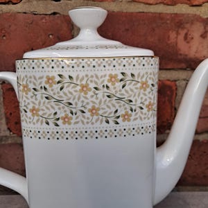 May include: White ceramic coffee pot with a lid and a long spout. The pot features a decorative band with yellow flowers, green leaves, and a dotted pattern. The rim is trimmed with gold.