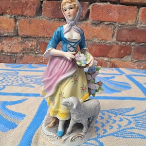 May include: A porcelain figurine of a person wearing a blue bodice, a pink apron, and a yellow skirt with floral patterns, holding a basket of flowers. A small grey sheep stands at the person's side, and the entire piece rests on a circular base.