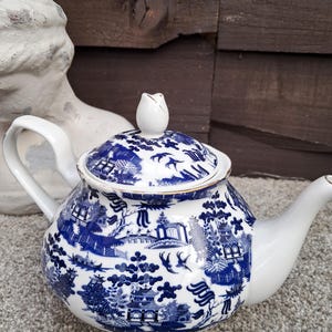 May include: A white ceramic teapot featuring a blue willow pattern with detailed illustrations of pagodas, bridges, and birds. The teapot has a rounded body, a curved handle, a spout, and a matching lid topped with a small white finial.