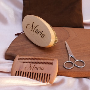 May include: A wooden beard comb and brush set with the name "Maria" engraved on each piece. The set is displayed on a wooden board with a pair of small scissors.