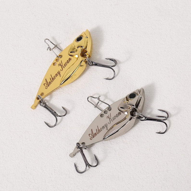 May include: Two gold and silver fishing lures with treble hooks. The lures are shaped like fish and have the name "Anthony Kwan" engraved on them.