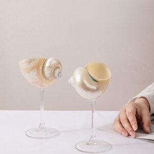 May include: Two decorative wine glasses, each with a seashell bowl and a clear stem. The shells display a pearlescent sheen with beige, white, and green hues. The glasses are on a white surface against a neutral backdrop.