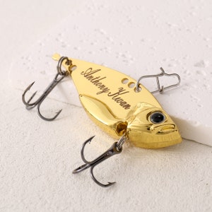 May include: Gold fishing lure with a black treble hook and a swivel. The lure is engraved with the name "Anthony Huaman".