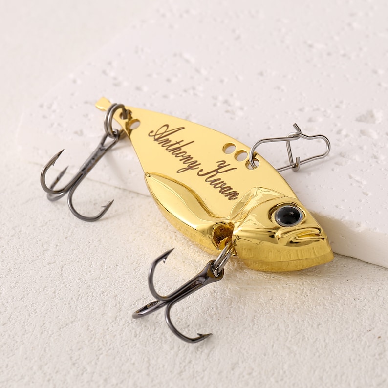 May include: Gold fishing lure with a black treble hook and a swivel. The lure is engraved with the name "Anthony Huaman".