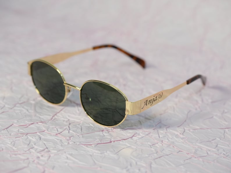 Puede incluir: Gafas de sol redondas doradas con lentes verde oscuro. Las gafas tienen el nombre "Anja" grabado en el lateral.