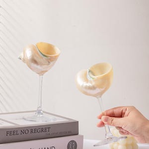May include: Two seashell-shaped wine glasses with clear stems and bases. The glasses have a pearlescent finish with a light yellow interior. One glass is held by a hand. The glasses sit on a stack of books. The books read "FEEL NO REGRET" and "HOLDER'S HAND."