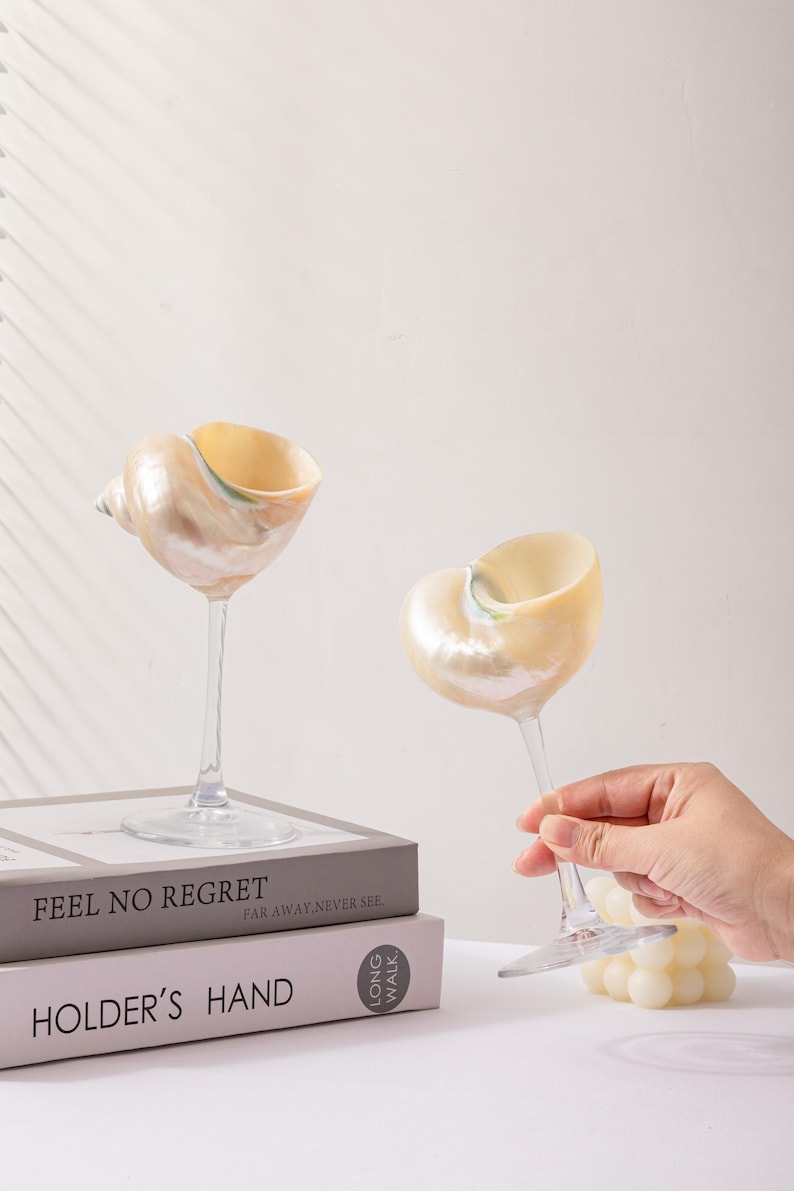 May include: Two seashell-shaped wine glasses with clear stems and bases. The glasses have a pearlescent finish with a light yellow interior. One glass is held by a hand. The glasses sit on a stack of books. The books read "FEEL NO REGRET" and "HOLDER'S HAND."