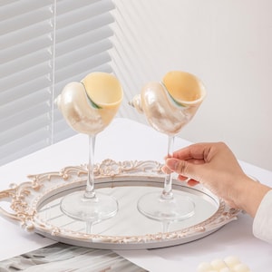 May include: Two seashell-shaped wine glasses with clear stems on a mirrored tray. The glasses have a pearlescent finish and are filled with a yellow liquid. A hand holds one glass. A white candle and decorative tray are also present.