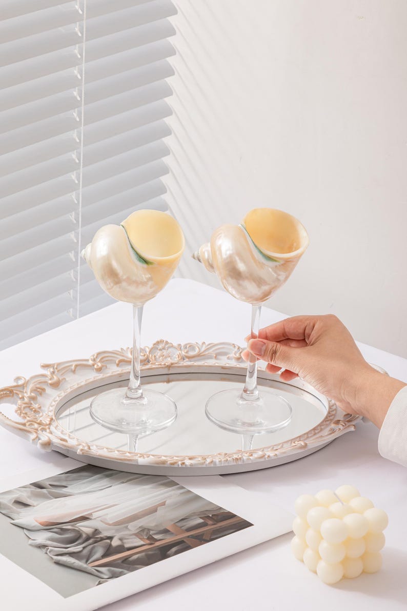 May include: Two seashell-shaped wine glasses with clear stems on a mirrored tray. The glasses have a pearlescent finish and are filled with a yellow liquid. A hand holds one glass. A white candle and decorative tray are also present.
